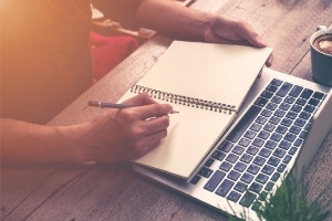 the hands of someone writing a synopsis in a journal; a laptop is also on the desk