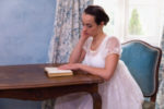 a regency era woman sitting at a desk reading a novel